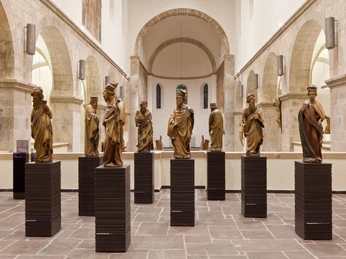 Museum Schnütgen Das Bild zeigt einen hell erleuchteten Museumsraum mit einer Reihe von hölzernen Skulpturen auf Sockeln.The picture shows a brightly lit museum room with a series of wooden sculptures on pedestals.