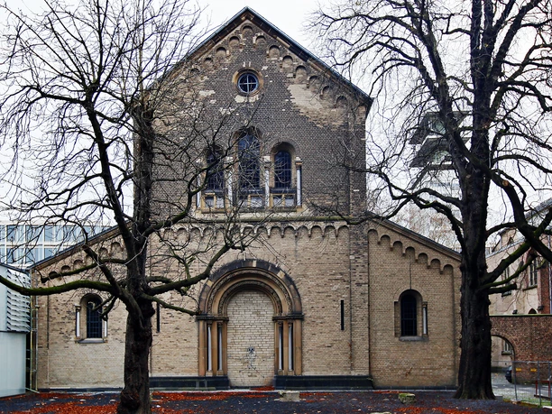 Sankt Cäcilien Das Bild zeigt die romanische Kirche Sankt Cäcilien in Köln. Die schlichte Backsteinfassade wird von zwei Bäumen flankiert, deren kahle Äste die Sicht auf das Bauwerk umrahmen.