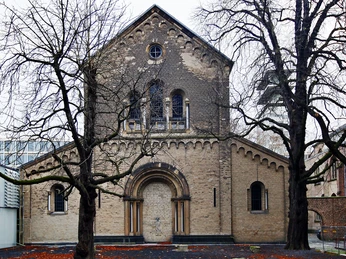 St. Cecilia Das Bild zeigt die romanische Kirche Sankt Cäcilien in Köln. Die schlichte Backsteinfassade wird von zwei Bäumen flankiert, deren kahle Äste die Sicht auf das Bauwerk umrahmen. The picture shows the Romanesque church of St. Cäcilien in Cologne. The simple brick façade is flanked by two trees whose bare branches frame the view of the building.
