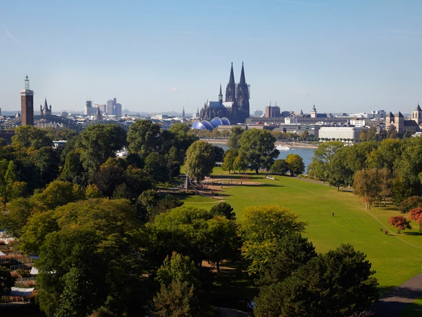 Rheinpark Das Bild zeigt den Kölner Rheinpark mit seiner weitläufigen Grünfläche. Im Hintergrund erheben sich majestätisch der Kölner Dom und die Altstadt, flankiert von der Hohenzollernbrücke. Bäume und Büsche säumen die Wiesen.