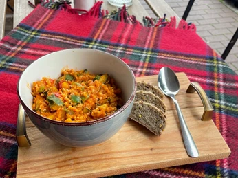 Mittagstisch bei PURPRODUKT.jpg Rustikale Mahlzeit: Eintopf in Schüssel, serviert auf Holzteller mit Brot, auf kariertem Tuch draußen.