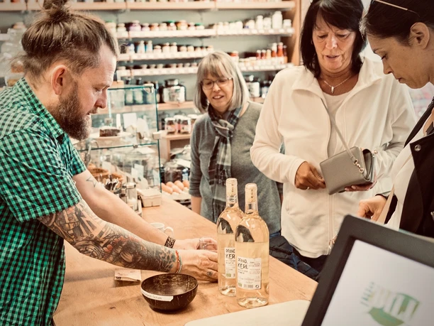 Ein tätowierter Mann präsentiert im Geschäft einer Gruppe Frauen drei Flaschen eines Getränks.
