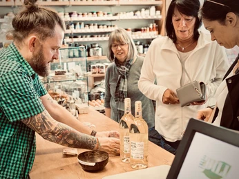 Service bei PURPRODUKT Ein tätowierter Mann präsentiert im Geschäft einer Gruppe Frauen drei Flaschen eines Getränks.