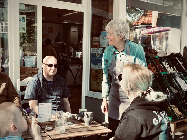 Außenplätze bei PURPRODUKT Menschen genießen Kaffee an Holztischen vor einem Café mit großer Fensterfront bei PURPRODUKT.