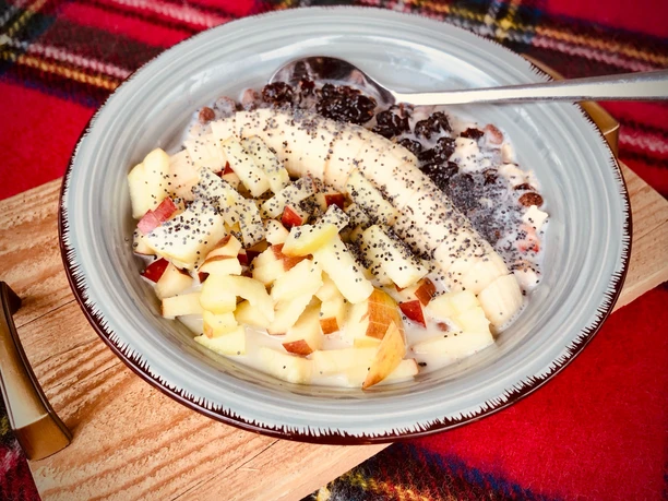 Eine Schale Müsli mit Bananen, Apfelstückchen, Mohn und Rosinen auf einem bunten Tisch.