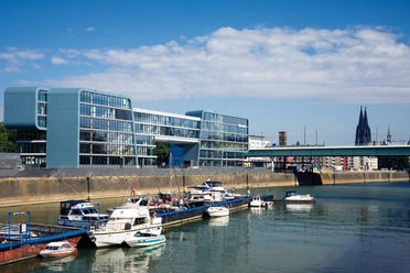 Rheinauhafen Modern office building on the waterfront with boats in the foreground, blue sky and Cologne Cathedral in the background.
