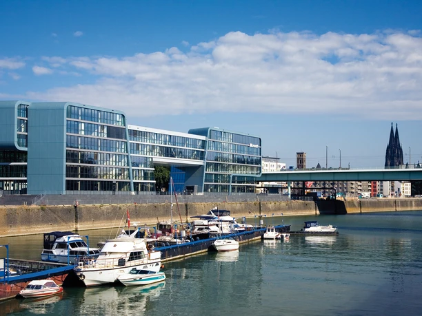 Rheinauhafen Modern office building on the waterfront with boats in the foreground, blue sky and Cologne Cathedral in the background.