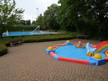 Gartenhallenbad Babybecken Buntes Babybecken mit Rutsche im Gartenhallenbad, umgeben von grünen Bäumen und Sitzbänken.