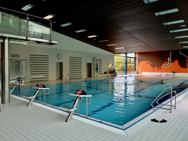 Schwimmhalle mit großem Becken, Rettungssteg und Leiter, helles Interieur mit bunter Wandgestaltung.