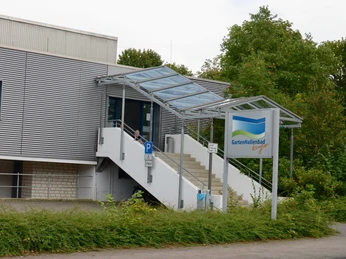 Eingangsbereich des Gartenhallenbads mit überdachter Treppe, umgeben von Grün und Parkplatz.