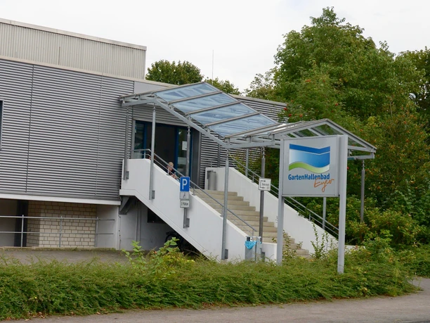 Eingangsbereich des Gartenhallenbads mit überdachter Treppe, umgeben von Grün und Parkplatz.
