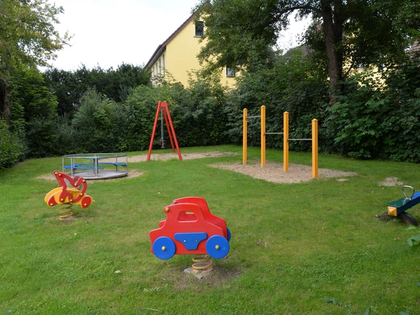 Spielplatz mit Wippe in Tierform, Klettergerüst, farbigen Stangen und umliegenden Bäumen.