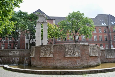 Römerbrunnen Eine ovale Brunnenanlage mit reliefverzierten, rötlichen Backsteinwänden, flankiert von Bäumen und einem Gebäude mit roten Ziegelwänden im Hintergrund.