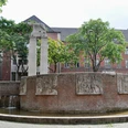 Römerbrunnen Eine ovale Brunnenanlage mit reliefverzierten, rötlichen Backsteinwänden, flankiert von Bäumen und einem Gebäude mit roten Ziegelwänden im Hintergrund.An oval fountain with reddish brick walls decorated in relief, flanked by trees and a building with red brick walls in the background.