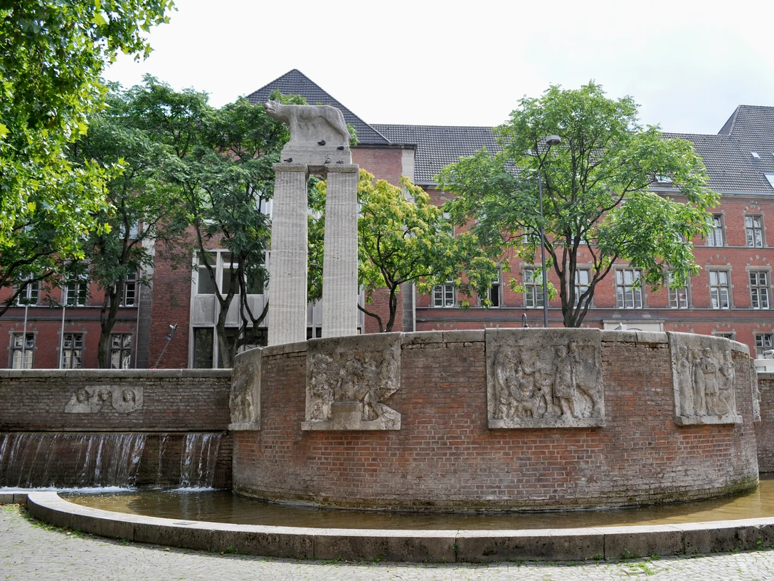 Roman fountain Eine ovale Brunnenanlage mit reliefverzierten, rötlichen Backsteinwänden, flankiert von Bäumen und einem Gebäude mit roten Ziegelwänden im Hintergrund.An oval fountain with reddish brick walls decorated in relief, flanked by trees and a building with red brick walls in the background.