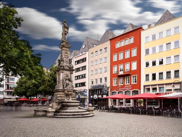 Jan-von-Werth Denkmal auf dem Alter Markt Ein lebhafter Platz in der Kölner Altstadt: Bunte Fassaden, Cafés mit roten Sonnenschirmen, zentraler Brunnen.