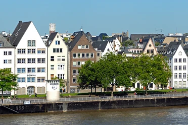 Pegel Köln Reihen schiefer Fachwerkhäuser mit spitzen Dächern säumen das Rheinufer unter strahlend blauem Himmel.