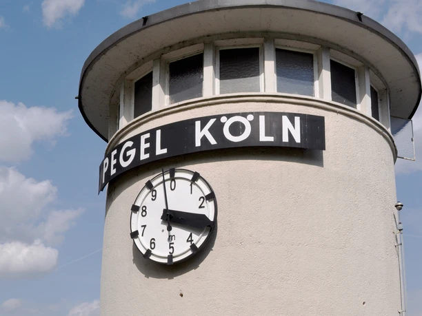 Pegel Köln Runder Pegelturm in Köln mit weißer Fassade und schwarzer Uhr, die drei Uhr zeigt.
