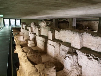 MiQua The picture shows the remains of the Roman governor's palace, the Praetorium, in Cologne. The old wall structures are partially preserved and illuminated by dim light, which emphasizes the historical atmosphere.