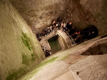 MiQua Das Bild zeigt die Mikwe in Köln, eine historische jüdische Badeanstalt. Von oben betrachtet, ist grünlich gealterte Steinmauer zu sehen, umgeben von Besucher*innen, die in den Schacht hinabsteigen.The picture shows the mikvah in Cologne, a historic Jewish bathing establishment. Viewed from above, a greenish aged stone wall can be seen, surrounded by visitors descending into the shaft.