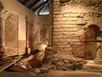 MiQua Im Bild sind die jahrhundertealten Mauern einer Mikwe in warmen, erdigen Farben zu sehen. Altertümliche Steinstrukturen und eine gedämpfte Beleuchtung schaffen eine meditative, ruhige Atmosphäre.The picture shows the centuries-old walls of a mikvah in warm, earthy colors. Ancient stone structures and subdued lighting create a meditative, calm atmosphere.