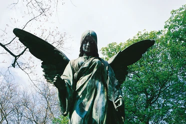 Melaten Friedhof Die majestätische Statue eines Engels mit weit geöffneten Flügeln erhebt sich erhaben vor einem Hintergrund aus blühendem Frühlingslaub und einem klaren Himmel.