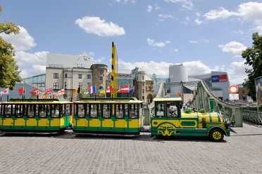 Wolters Bimmelbahn Eine gelb-grüne Bimmelbahn fährt über Kopfsteinpflaster, im Hintergrund moderne und historische Gebäude.