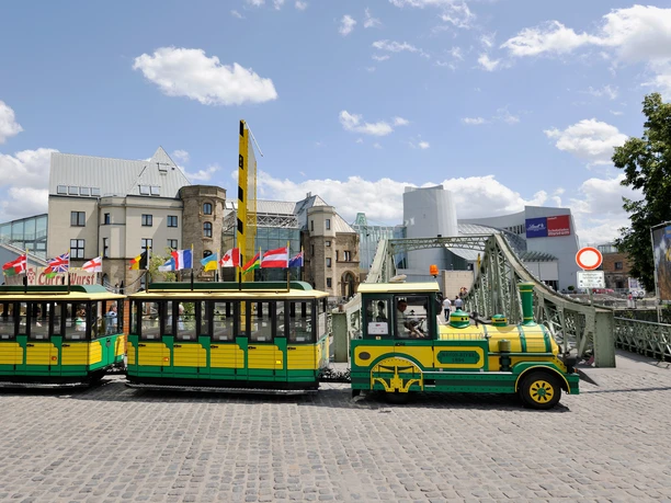 Wolters Bimmelbahn A yellow and green train runs over cobblestones, with modern and historic buildings in the background.