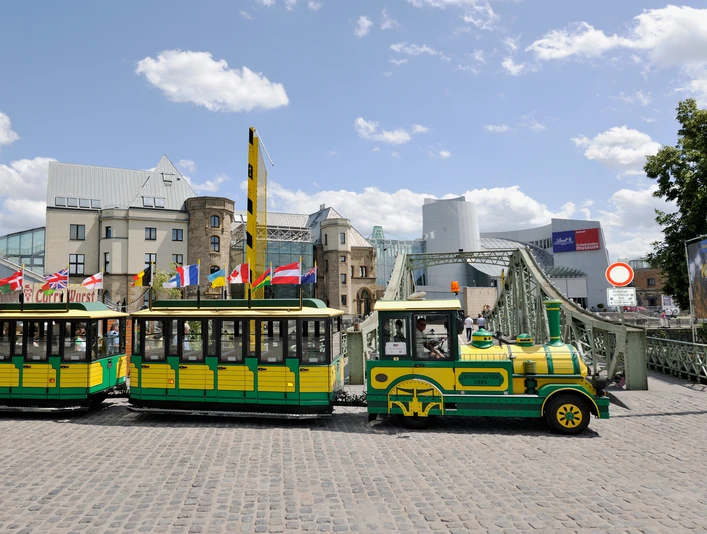 Wolters Bimmelbahn Eine gelb-grüne Bimmelbahn fährt über Kopfsteinpflaster, im Hintergrund moderne und historische Gebäude.A yellow and green train runs over cobblestones, with modern and historic buildings in the background.