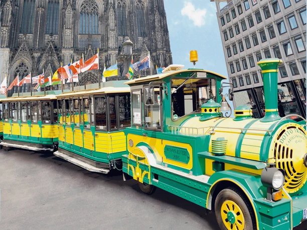 Wolters Bimmelbahn A yellow and green train stands in front of the imposing Cologne Cathedral, flanked by a modern building.