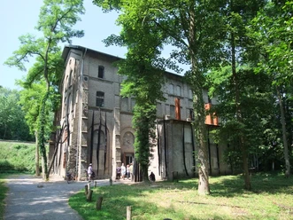 The Augustaschacht Memorial, surrounded by trees, with a historic brick building in the background.