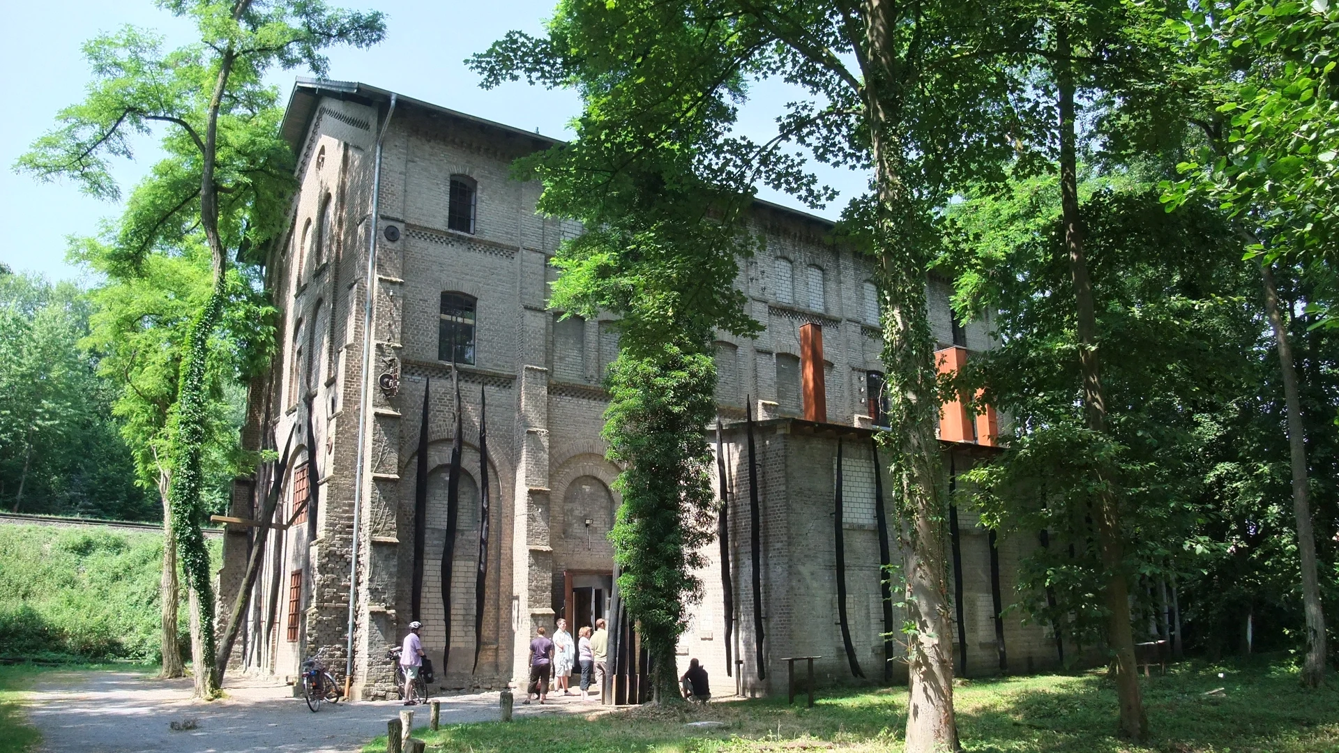 The Augustaschacht Memorial, surrounded by trees, with a historic brick building in the background.