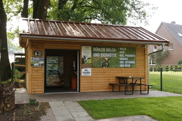 Rühler Milch- und Eierhütte Kleine Verkaufshütte aus Holz mit Milchautomat, Werbetafel und Sitzbank unter Bäumen im Grünen.