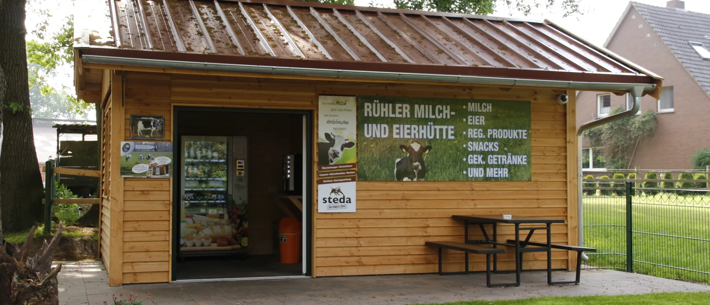 Rühler Milch- und Eierhütte Kleine Verkaufshütte aus Holz mit Milchautomat, Werbetafel und Sitzbank unter Bäumen im Grünen.