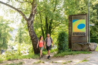Ein Pärchen in Wanderklamotten spaziert an Bäumen und einer Infotafel mit der Aufschrift "Eifelsteig" vorbei.