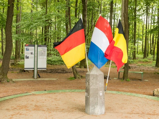 Dreiländerpunkt, an dem die Grenzen von Deutschland, Belgien und den Niederlanden aufeinandertreffen. Drei Flaggen repräsentieren die Länder und sind um einen Stein angeordnet, der als Symbol für den Grenzpunkt dient. Im Hintergrund ist ein Wald und touristische Informationstafeln.