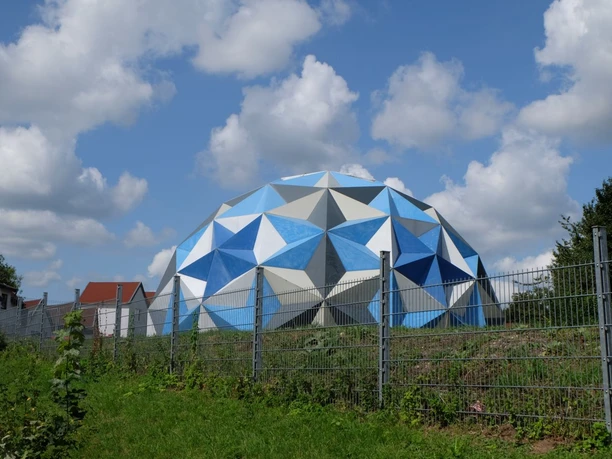 Campus Melaten Eine auffällige Kuppel mit blauen und weißen geometrischen Mustern erhebt sich hinter einem Metallzaun unter blauem Himmel.