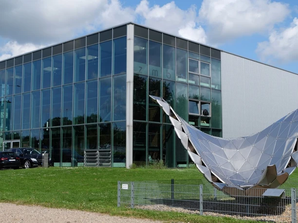 Modernes Gebäude mit Glasfassade und Skulptur im Vordergrund.