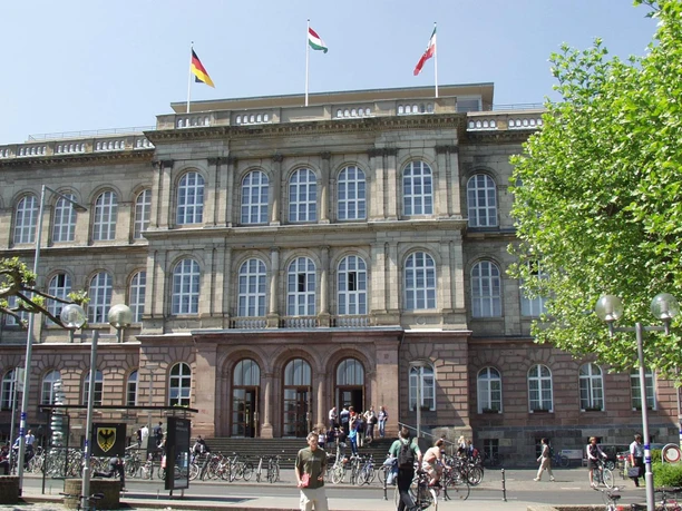 Historisches Bauwerk mit symmetrischen Fensterreihen, drei Eingangstüren, Fahnen auf dem Dach und einer Fassade in hellem Grau. Vor dem Gebäude erstreckt sich ein Platz, über den Menschen spazieren.