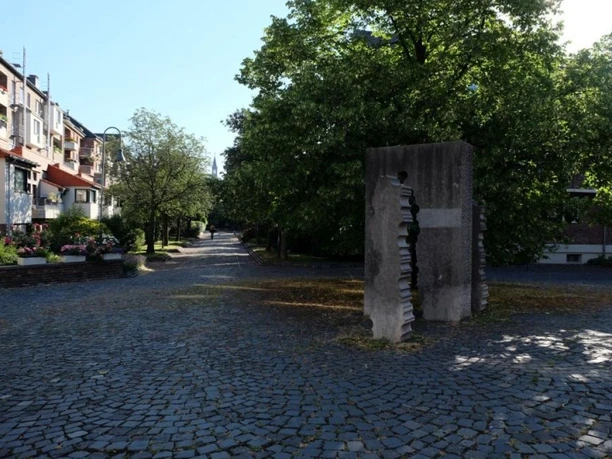 Ruhige Wohngegend. Ein Denkmal aus Stein steht auf gepflastertem Platz, unter Bäumen und neben blühenden Sträuchern.