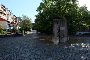 Hermann-Heusch-Platz Ruhige Wohngegend. Ein Denkmal aus Stein steht auf gepflastertem Platz, unter Bäumen und neben blühenden Sträuchern.