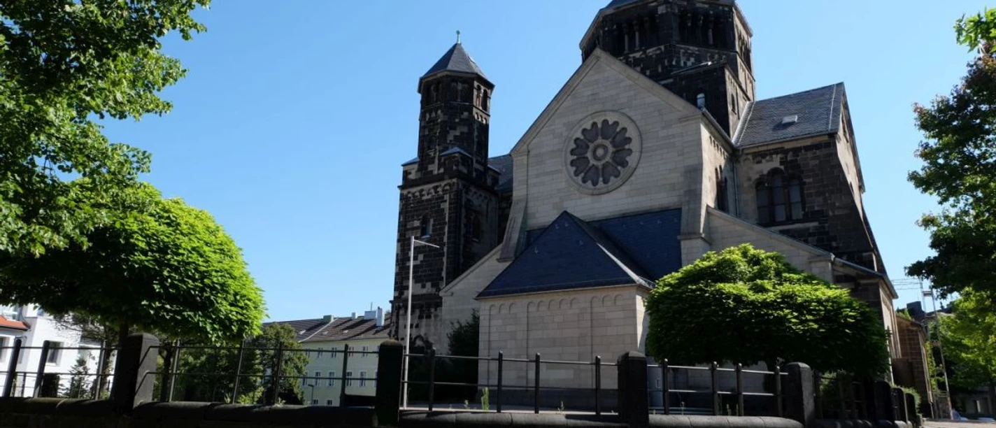 Romanische Kirche mit runder Rosette und markanten Türmen, an einem sonnigen Tag, umgeben von Bäumen und gepflegten Grünflächen.