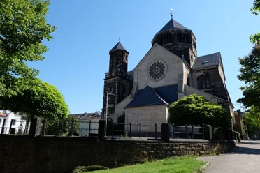 Herz-Jesu Kirche Romanische Kirche mit runder Rosette und markanten Türmen, an einem sonnigen Tag, umgeben von Bäumen und gepflegten Grünflächen.