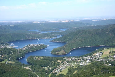Der Rursee schlängelt sich mal sehr breit, mal etwas schmaler, durch hügelliges, dicht bewaldetes Land. Von oben sind kleine Boote auf dem Wasser zu erkennen.