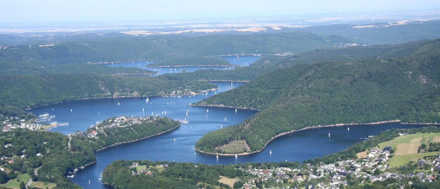 Eifel Der Rursee schlängelt sich mal sehr breit, mal etwas schmaler, durch hügelliges, dicht bewaldetes Land. Von oben sind kleine Boote auf dem Wasser zu erkennen.