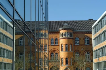 Blick an einer Glasfassade entlang auf ein historisches Gebäude mit ockerfarbenen Steinen.