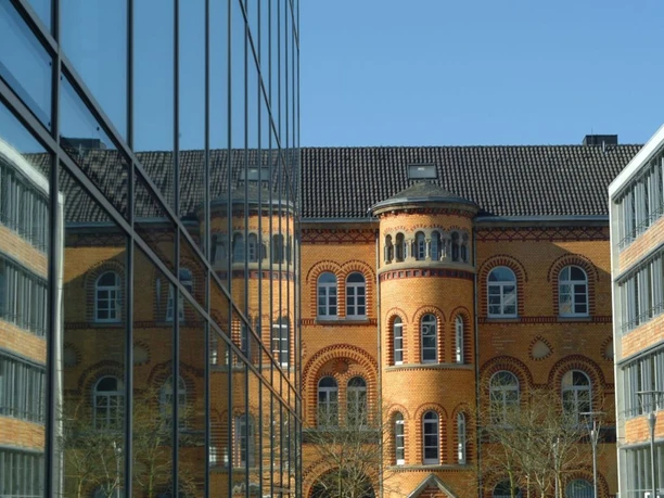 Kapuzinerkarree Blick an einer Glasfassade entlang auf ein historisches Gebäude mit ockerfarbenen Steinen.