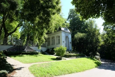 Kerstenscher Pavillon Weiß gestrichenes Gebäude im neoklassizistischen Stil im Sonnenlicht hinter einem gepflegten Vorgarten mit Bäumen.
