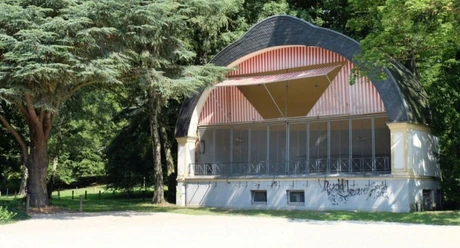 Konzertmuschel aus Holz mit gewölbtem Dach, umgeben von grünen Bäumen im Park.