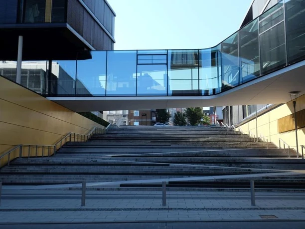 AachenMünchener Moderner Gebäudekomplex mit einer gläsernen Überführung, die zwei Gebäudeteile verbindet. Eine breite Treppe führt nach oben unter der Überführung hindurch..